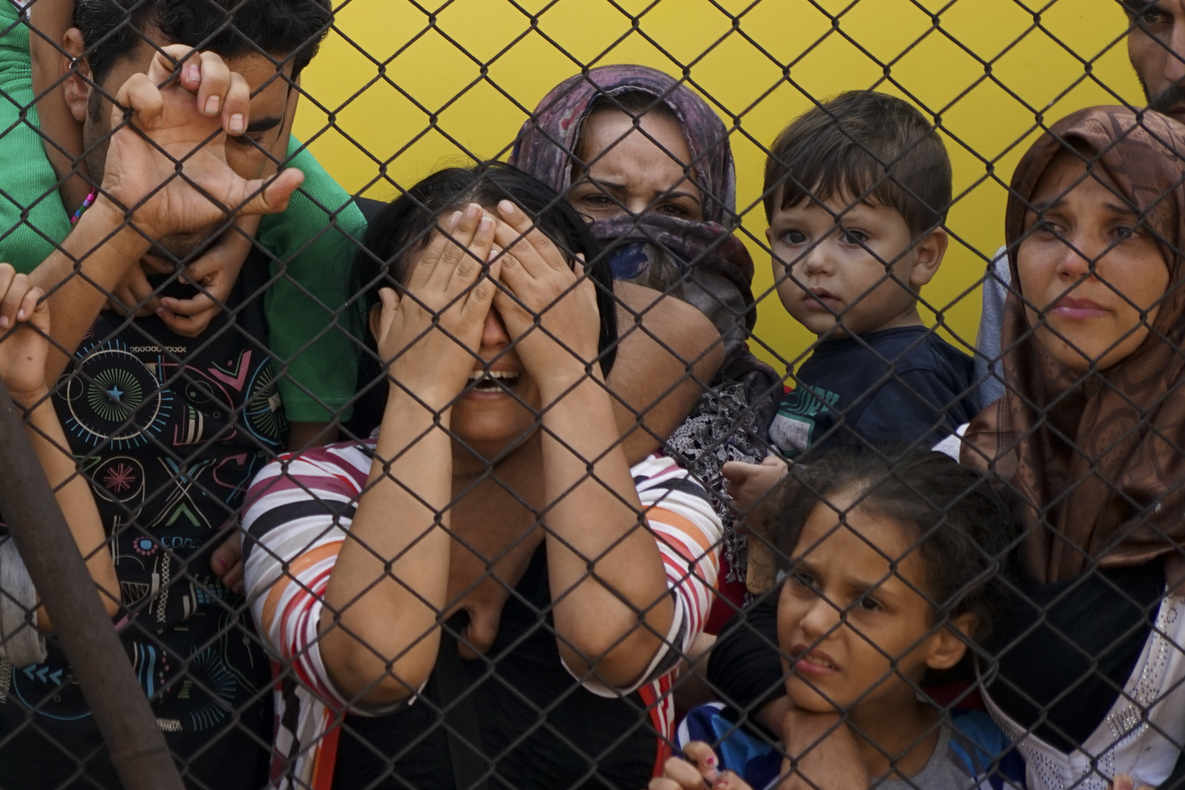 A group of children, women and a young man behind a chain link fence, one woman with hands over her eyes, wailing