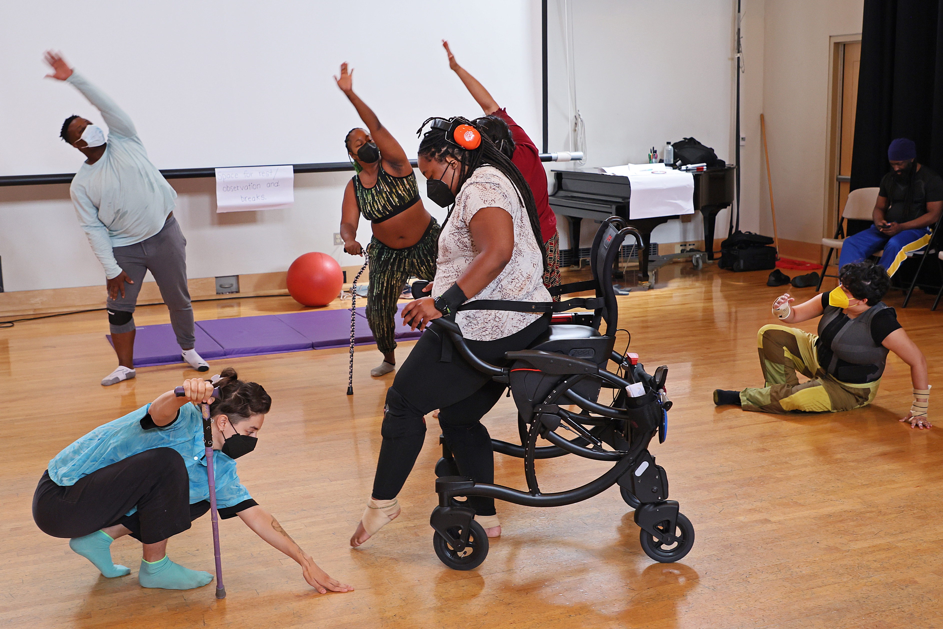 Six people in a dance studio. Some use mobility assistive devices, some are reaching their left arms over their heads, others are crouching or sitting on the floor.