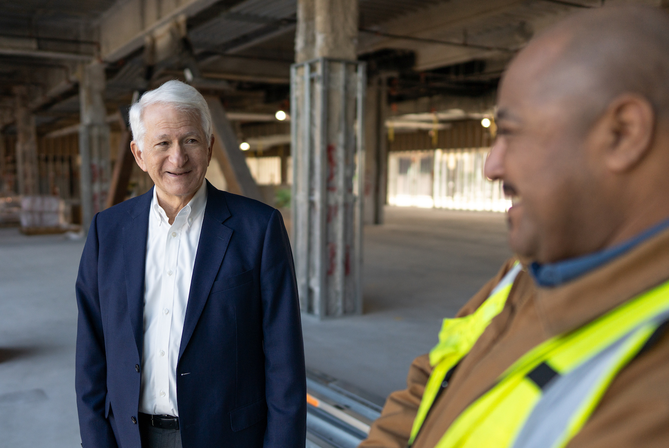 An older white man in a blazer, Gene Block, chats with a construction worker in a yellow vest at a construction site