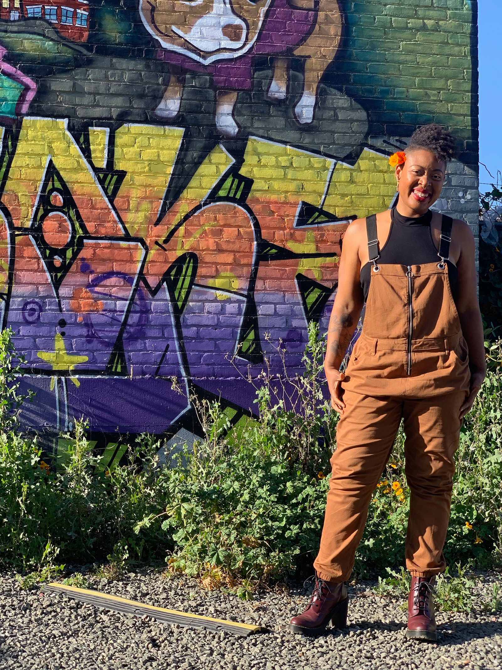 Ayana Omilade Flewellen stands in front of a wall with colorful murals outside, wearing overalls, smiling