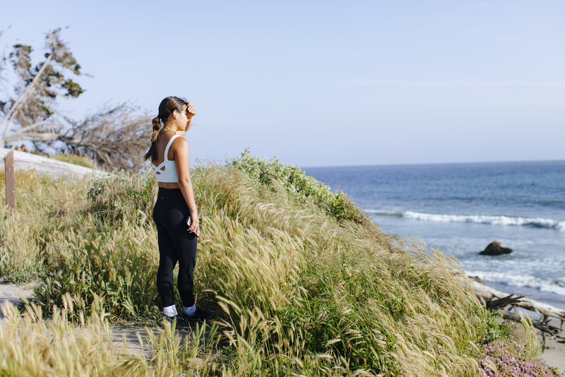 Young woman wearing workout attire shades her eyes with her hand while looking out over the ocean from a grassy bluff.