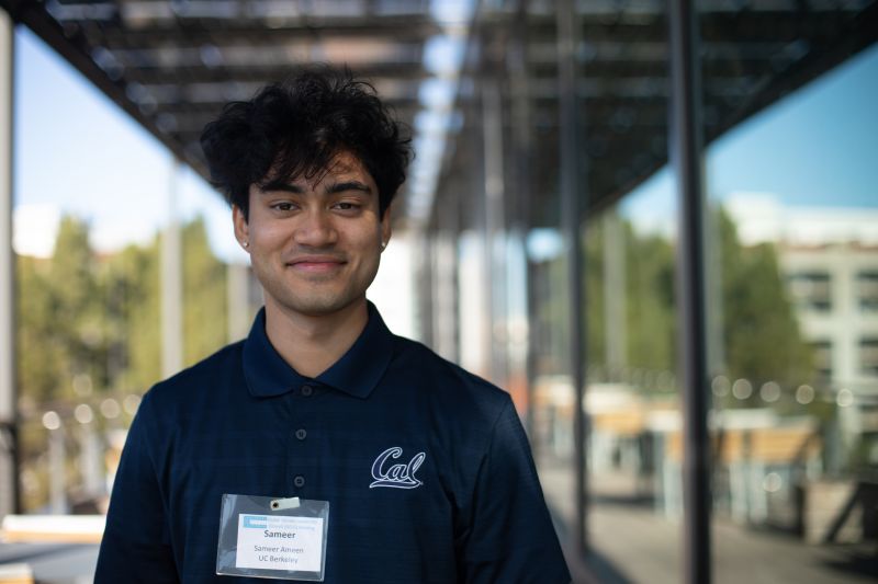 Sameer Ameen, wearing a Cal polo and name tag, smiles at the camera with a glass wall in the background reflecting a sunny day.