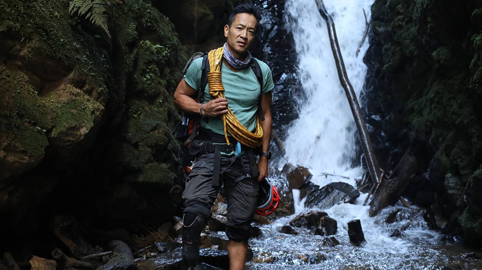 Albert Lin in outdoorsy gear stands in front of a waterfall