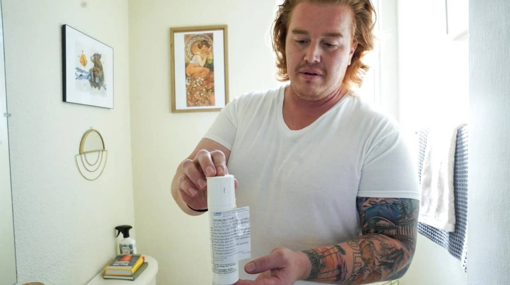 A man with arm tattoos in a white t-shirt holds a white plastic bottle, standing in a well lit room with white walls.