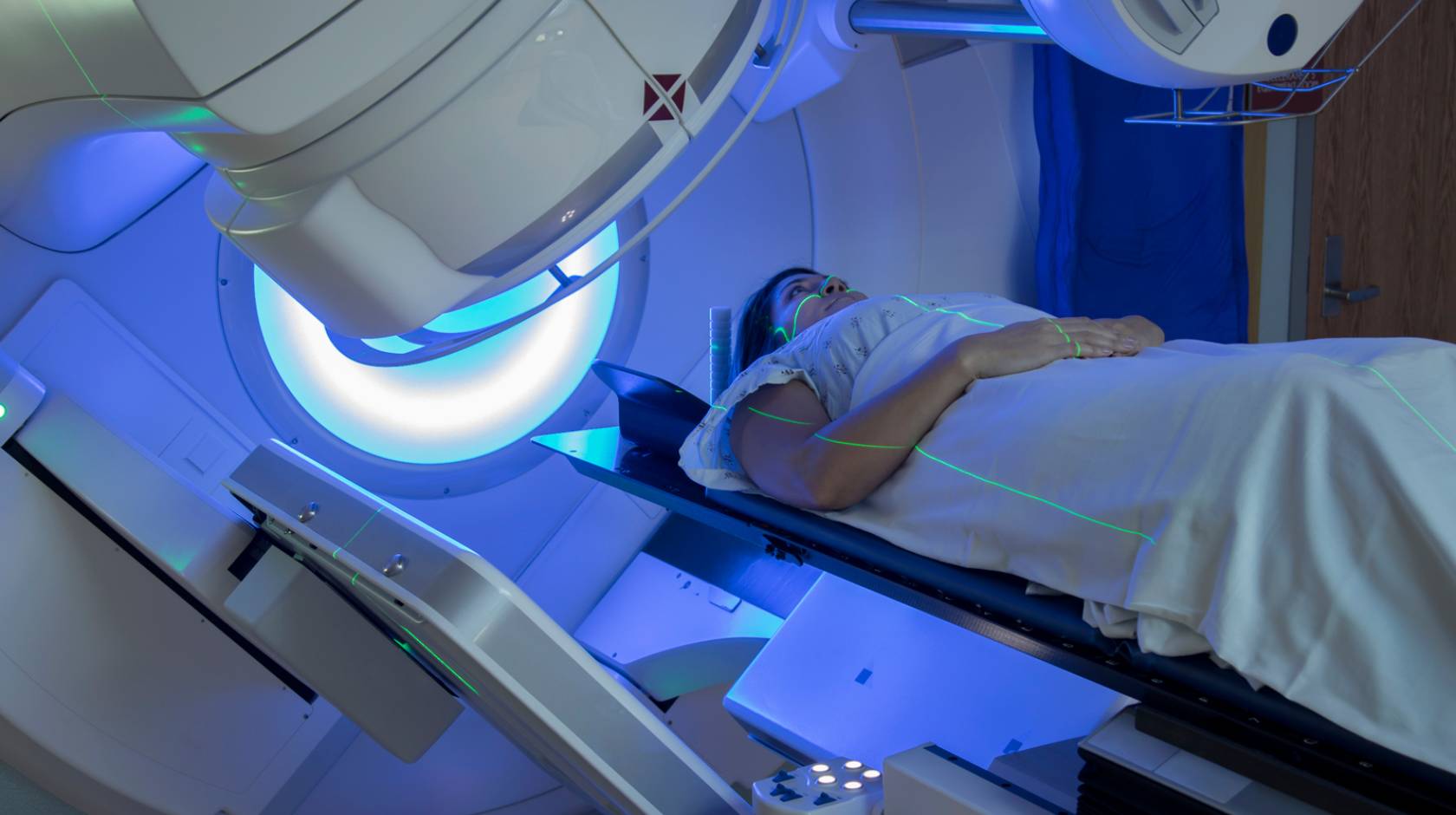 Woman receiving radiation therapy treatments for breast cancer under a machine with green lines of light on her face and body