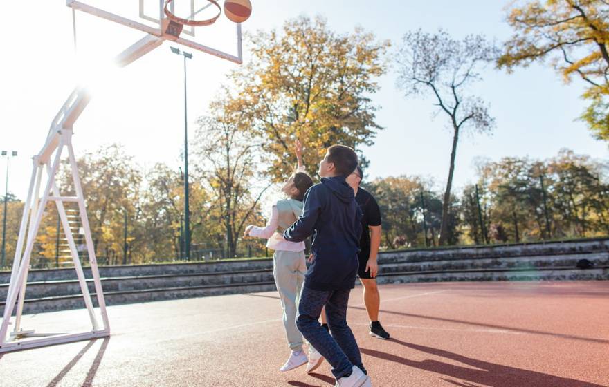 Two kids play outdoors on a basketball court with a parent
