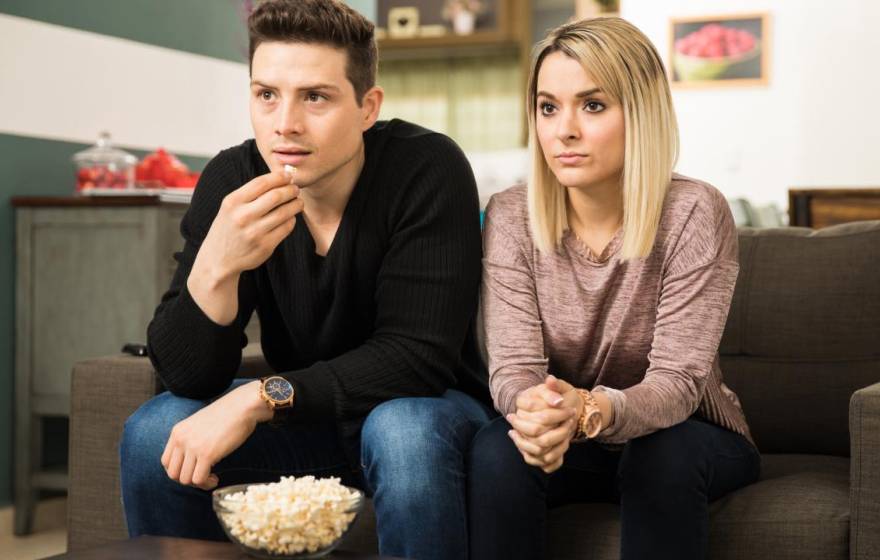 A white man and a white woman watching TV together, looking somber