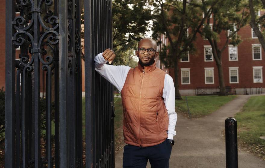 Young Black man in an orange vest and glasses standing against a gate on the Harvard campus