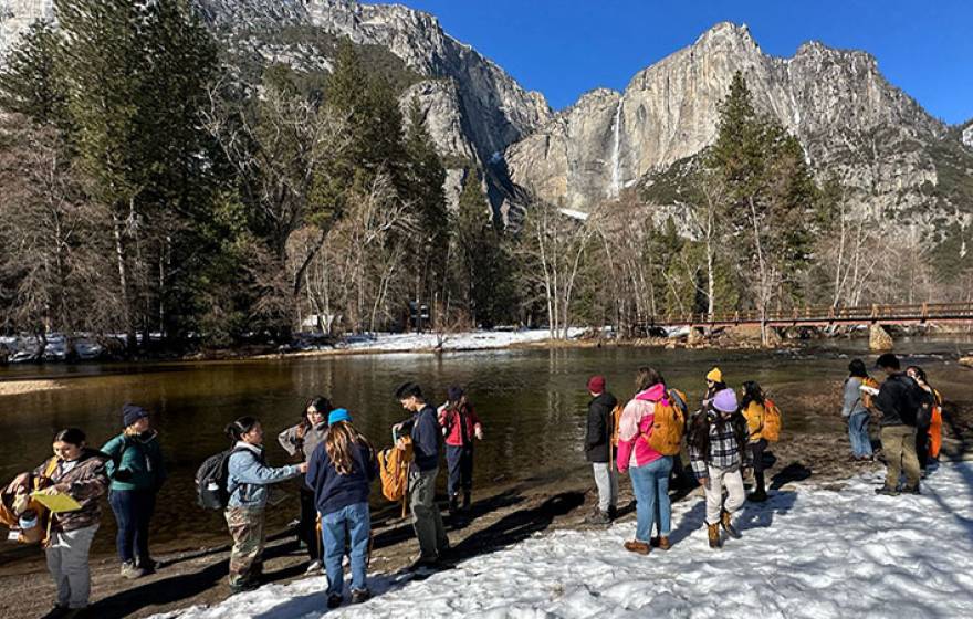 On a sunny day, a group of students gathers on the banks of a river, with snow on the ground and dramatic mountain peaks with a waterfall and forest in the background. 