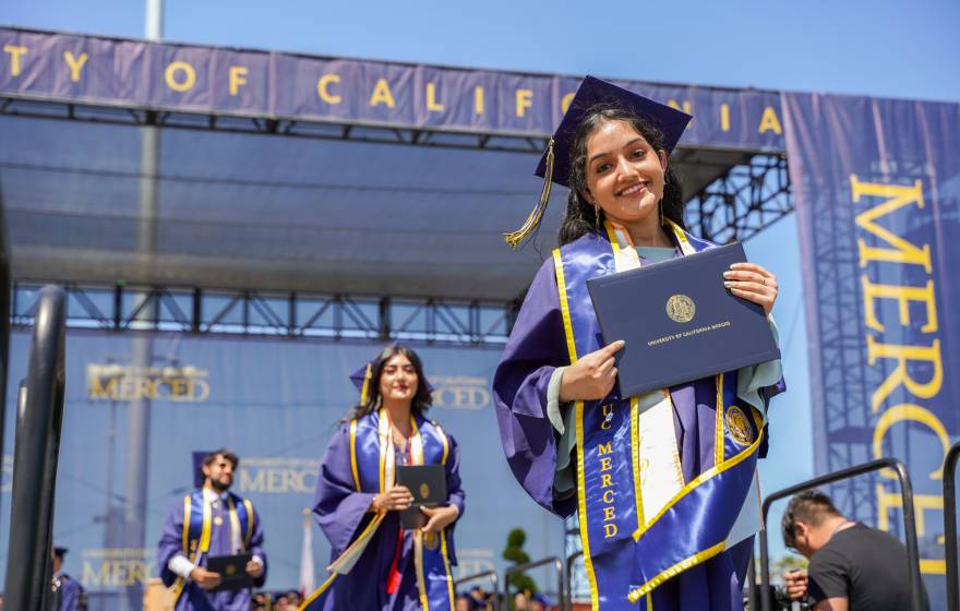 Three UC Merced graduates hold their diplomas as they leave the stage of graduation, smiling