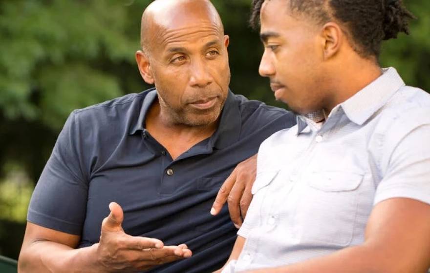 A father and his teen son sit on a park bench. The father is speaking intently to the son, who appears to be listening.