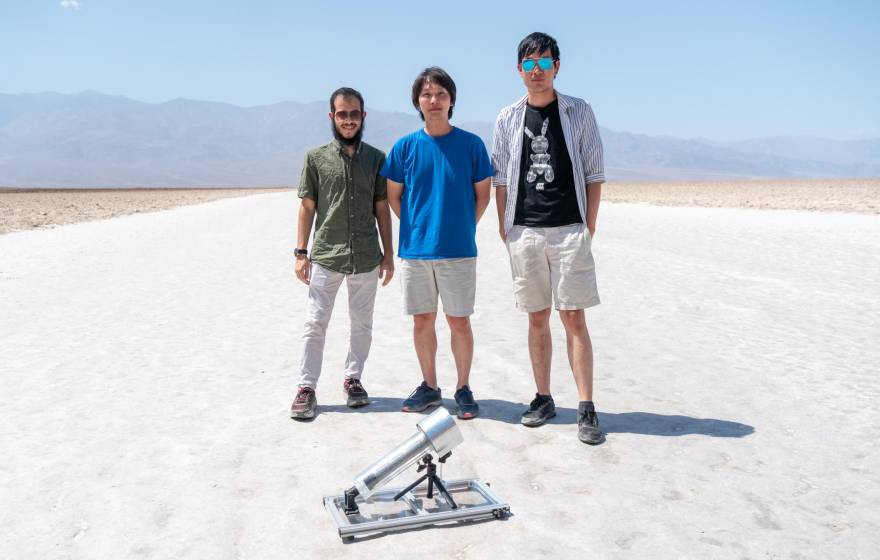 Three male students stand in white desert sands, behind a water harvester that looks like a microphone
