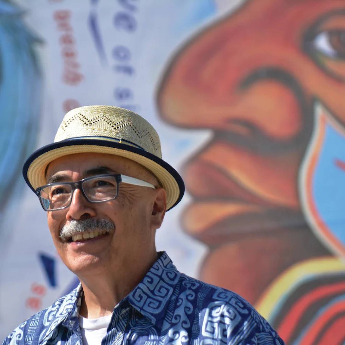 An older Latino man with a mustache, glasses and a jaunty hat in front of a colorful mural