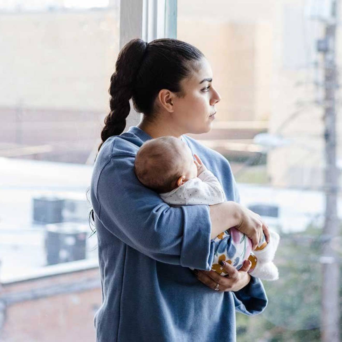 A woman with a long ponytail holds a baby and looks out the window with a trouble expression on her face