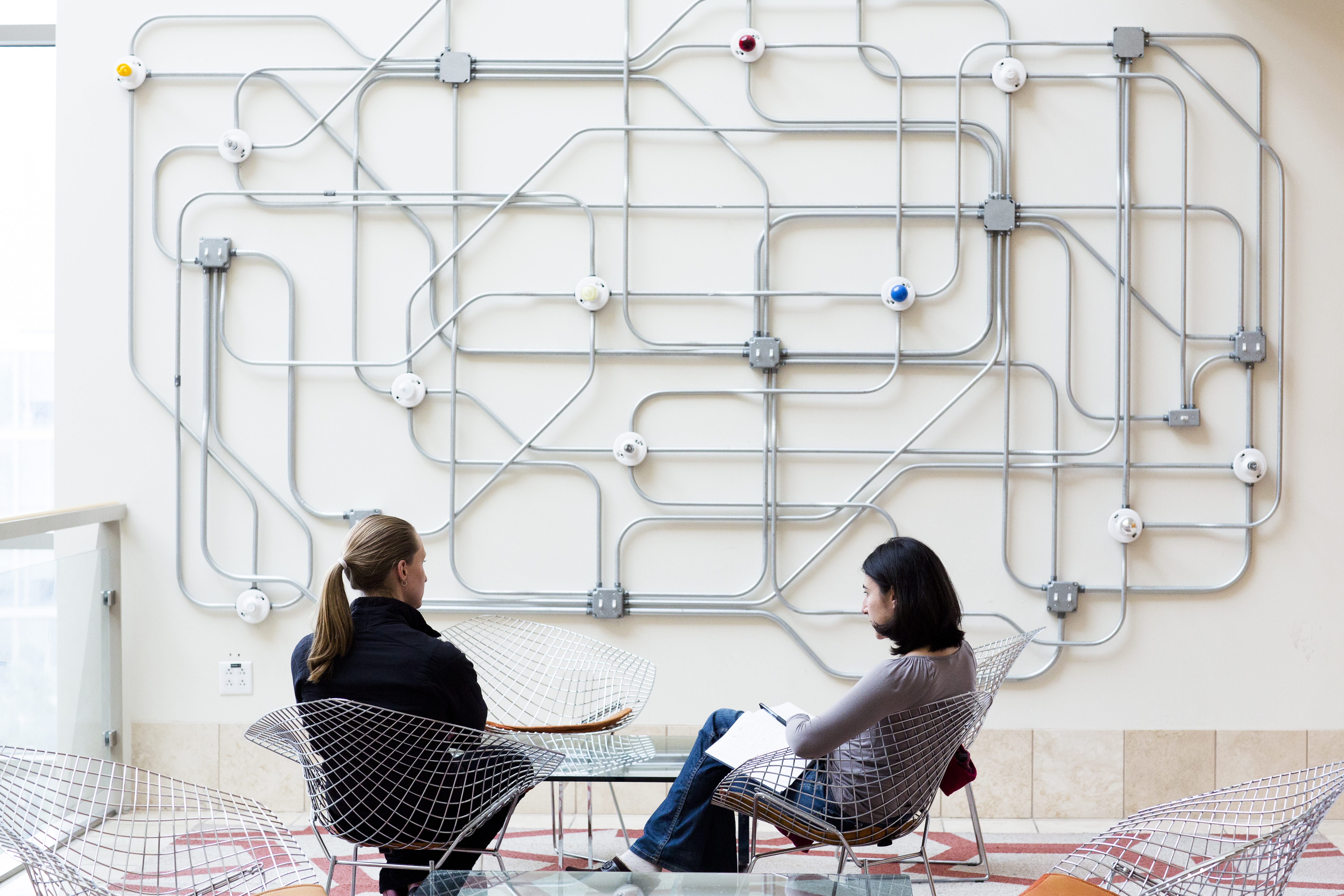 Two women in chairs talking in front of an interesting display of tubes