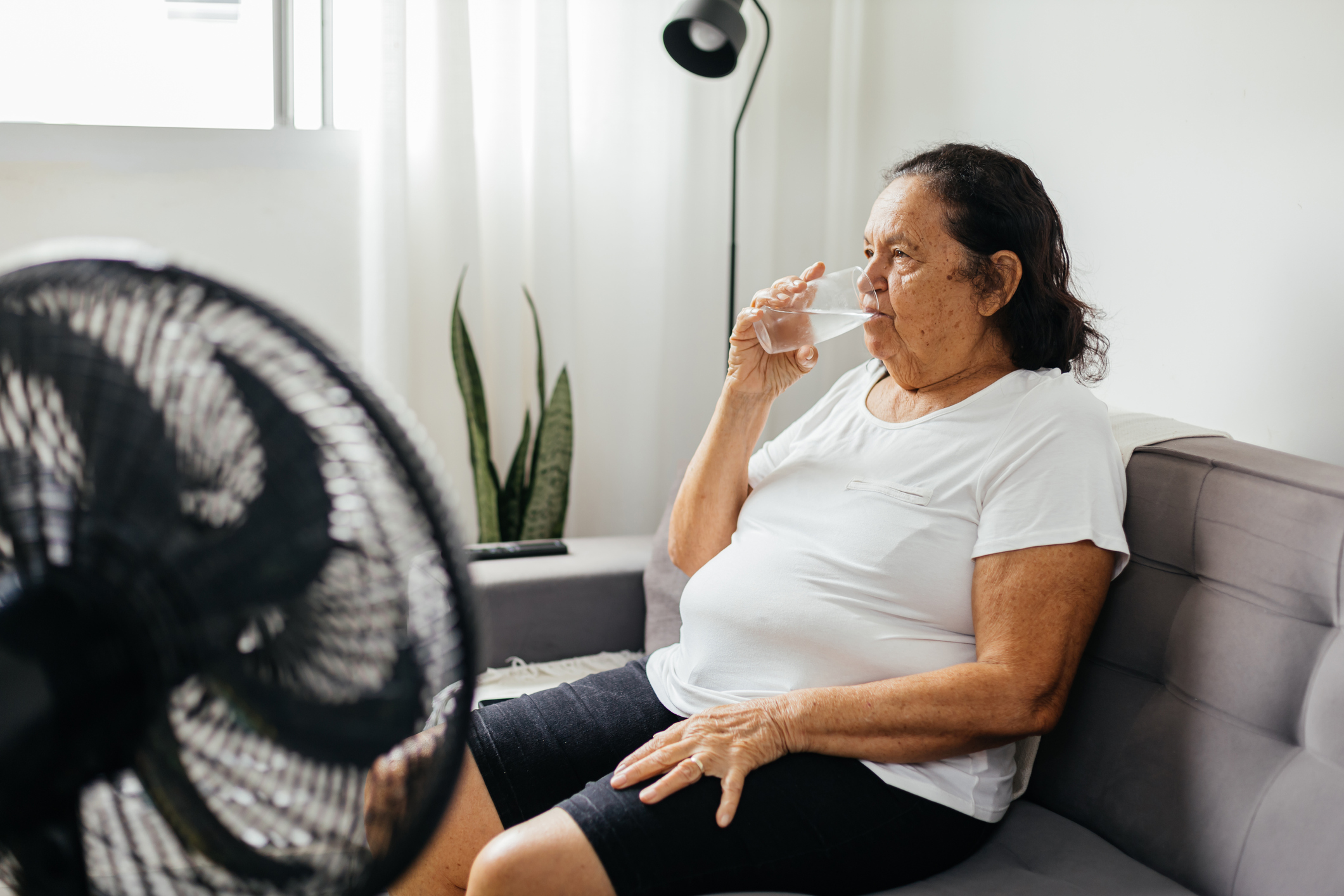 An older Latina woman drinking water in front of a fan inside
