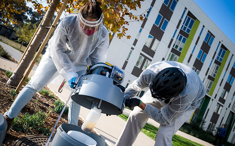 Two people in hazmat suits pulling wastewater samples out with a machine