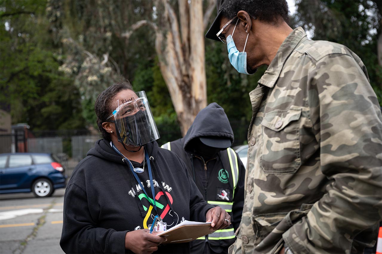A Black worker with a face shield and a clipboard talks to a Black man in camo