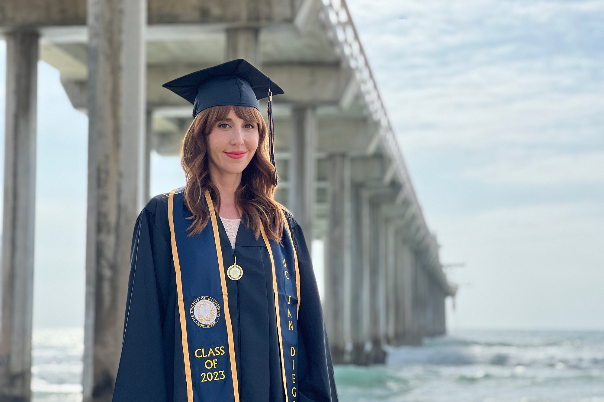 White woman in cap and gown at San Diego pier, ocean behind