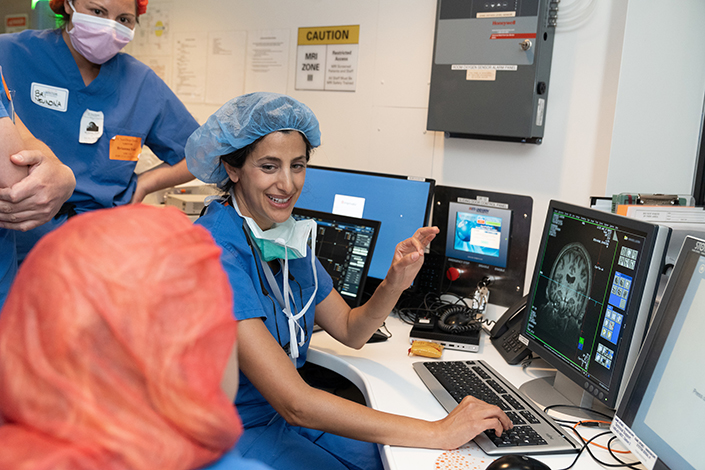 A woman surgeon in scrubs talking to three other people in scrubs, looking at a computer with a brain scan
