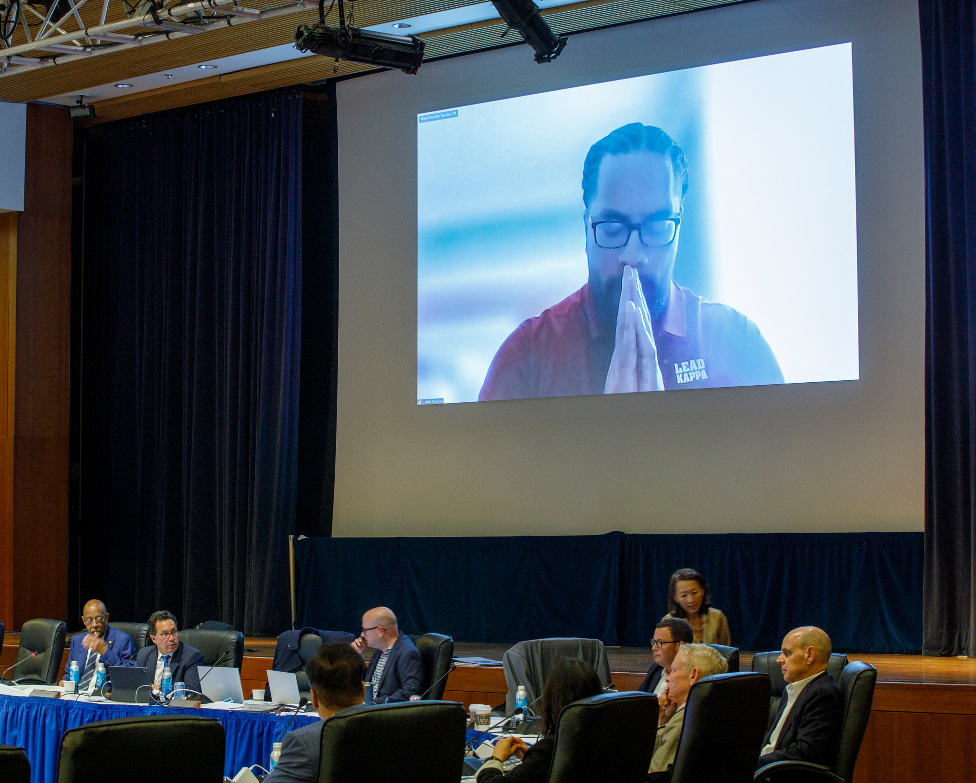 Abiel Malepeai, a young Samoan man with a beard and glasses, holds his hands clasped over his mouth, eyes lowered, while speaking on a projector screen to the Regents meeting