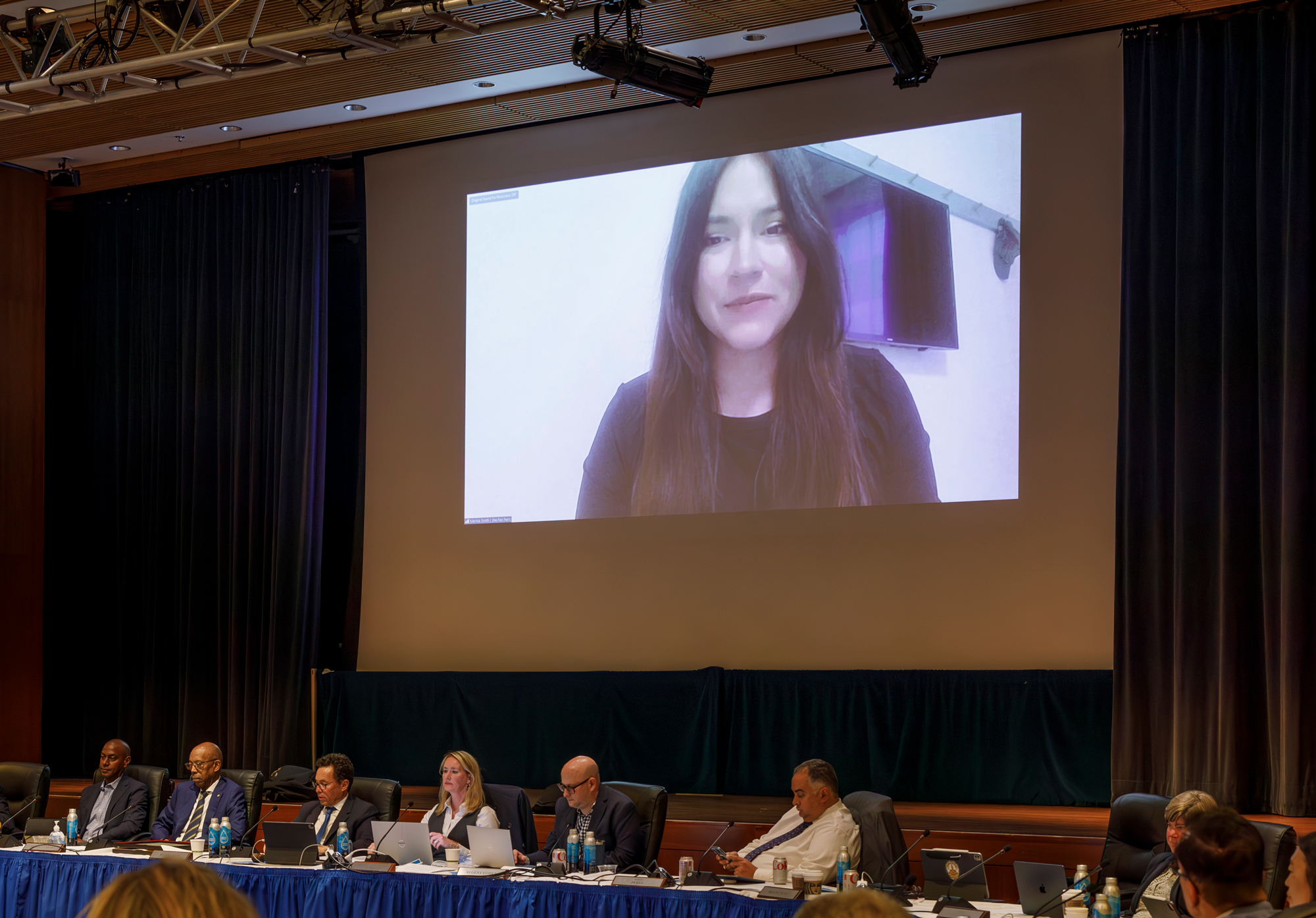 A young woman with dark long hair, Merina Smith, delivers remarks to the Regents from a projector screen via Zoom