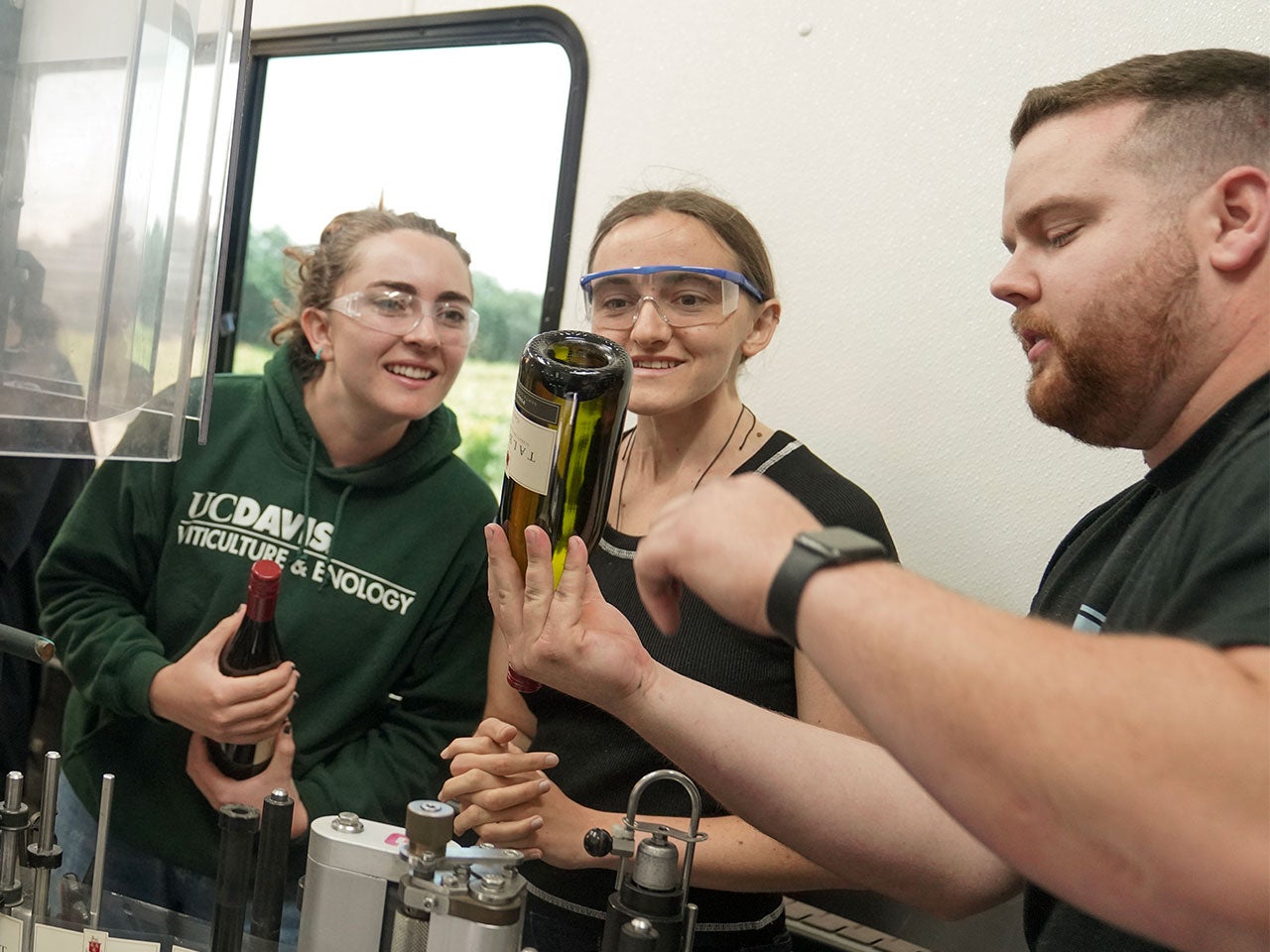 Two women wearing safety glasses watch as a man turns a wine bottle upside down, showing them something