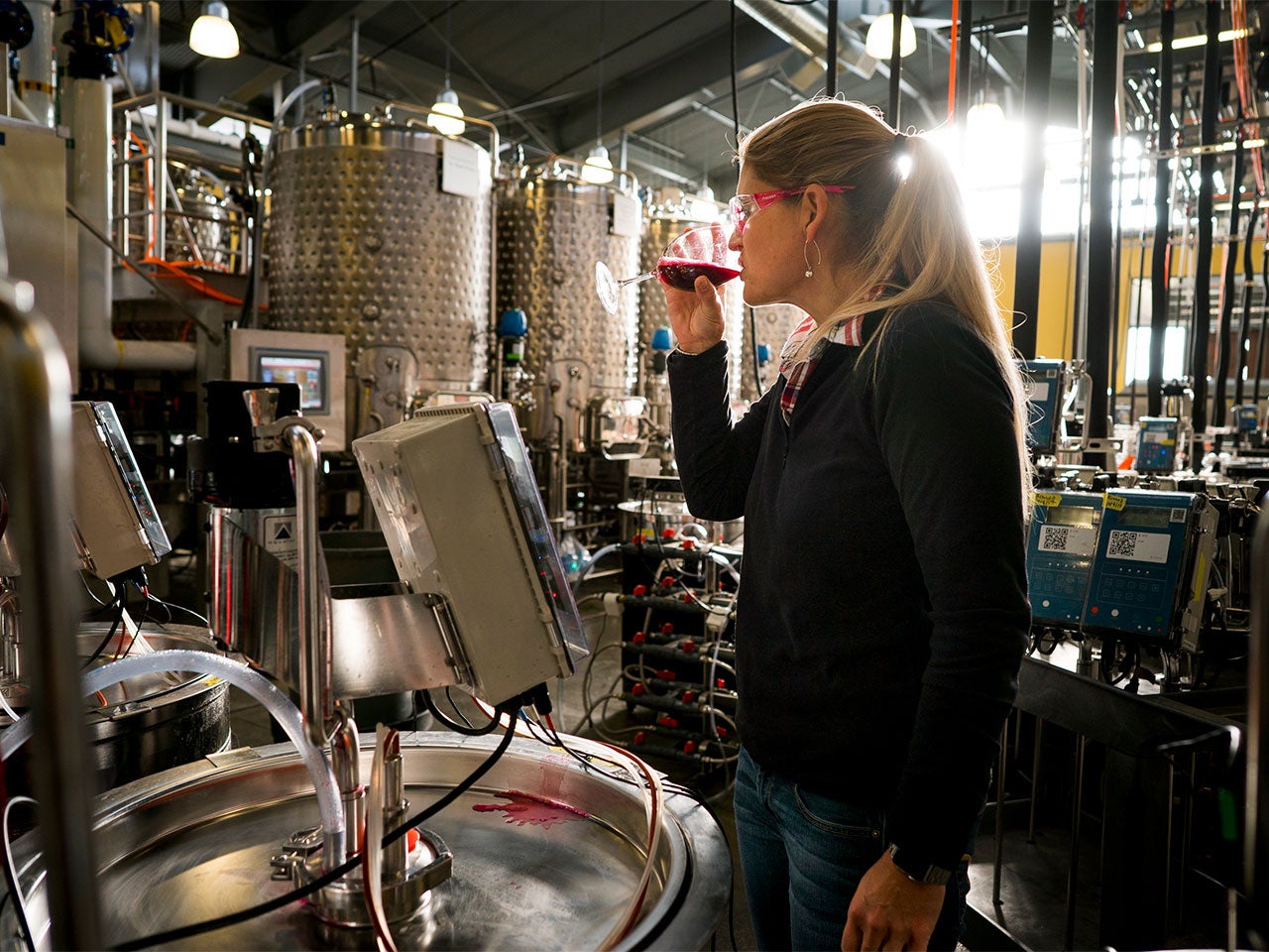 A blonde woman in safety glasses drinks a glass of wine surrounded by equipment