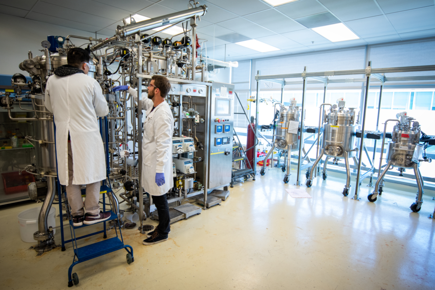 Two people in white lab coats and goggles work on metallic lab equipment