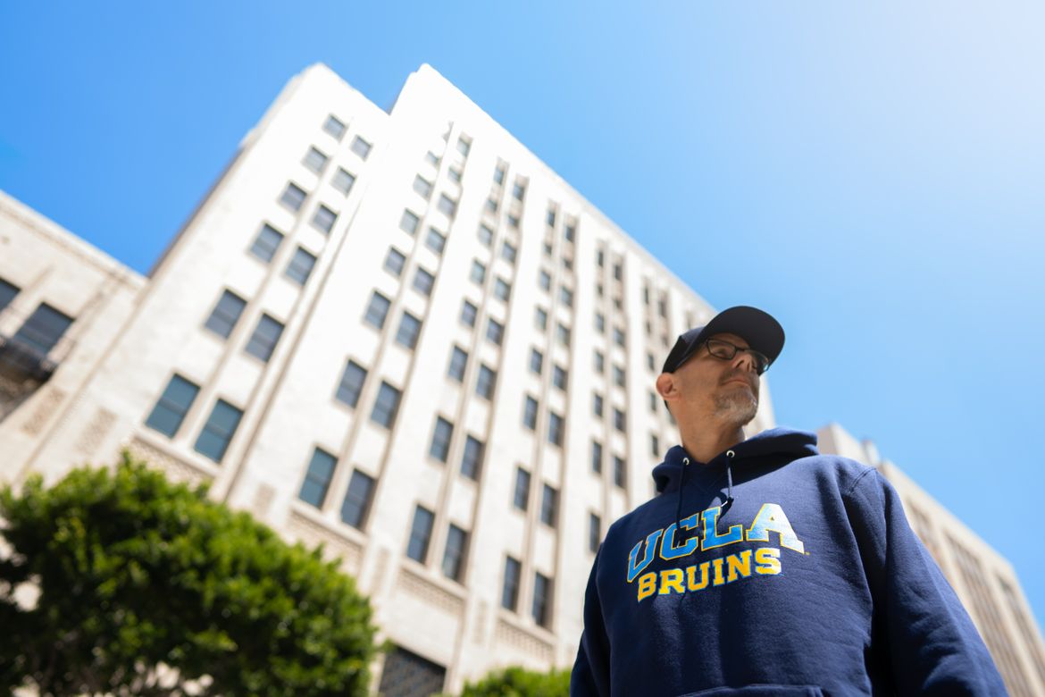 A man in a UCLA hooded sweatshirt in front of the Trust Building, a tall white building in Los Angeles