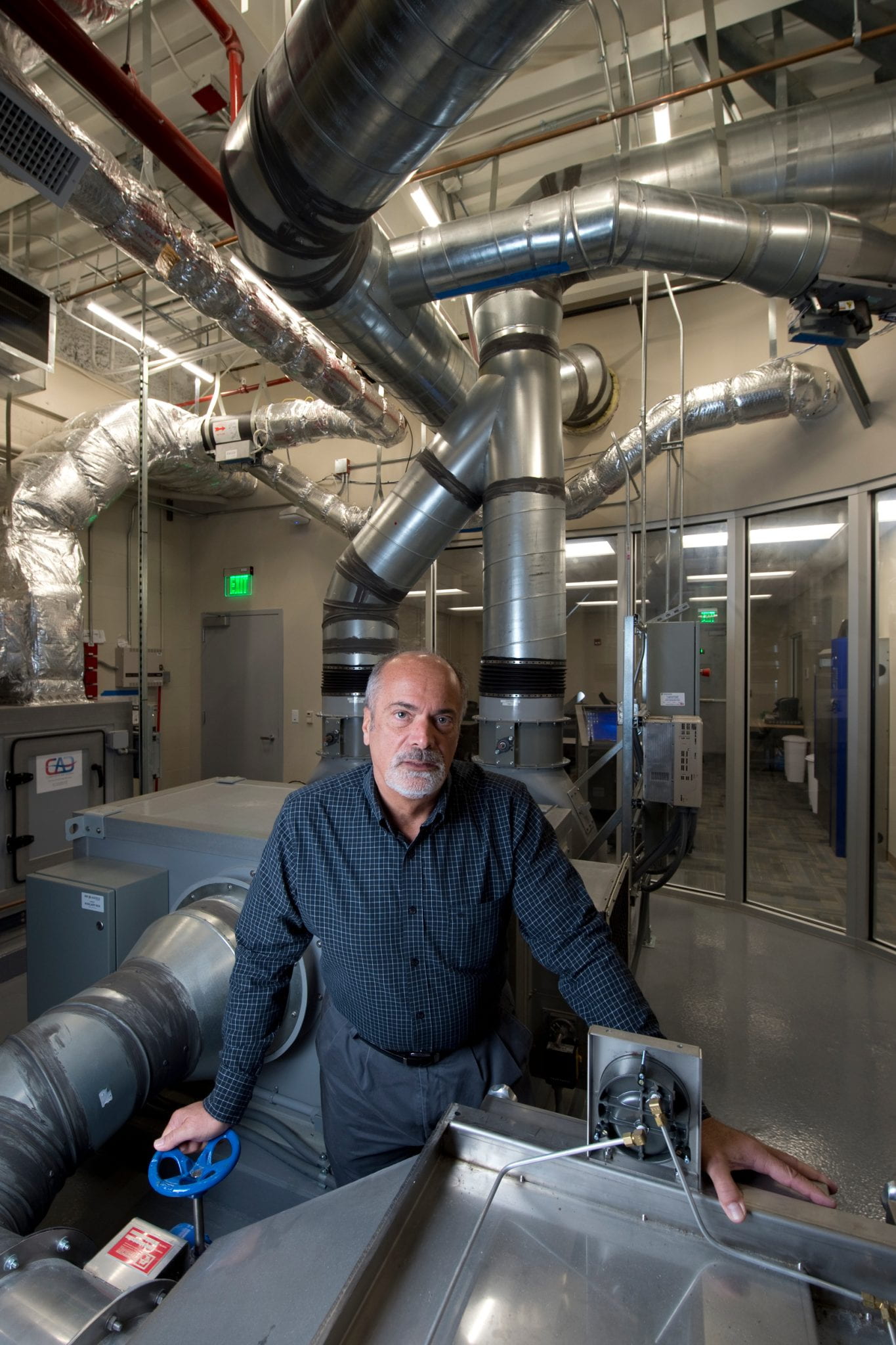 A man with a white goatee in a large lab environment with lots of ventilation around him