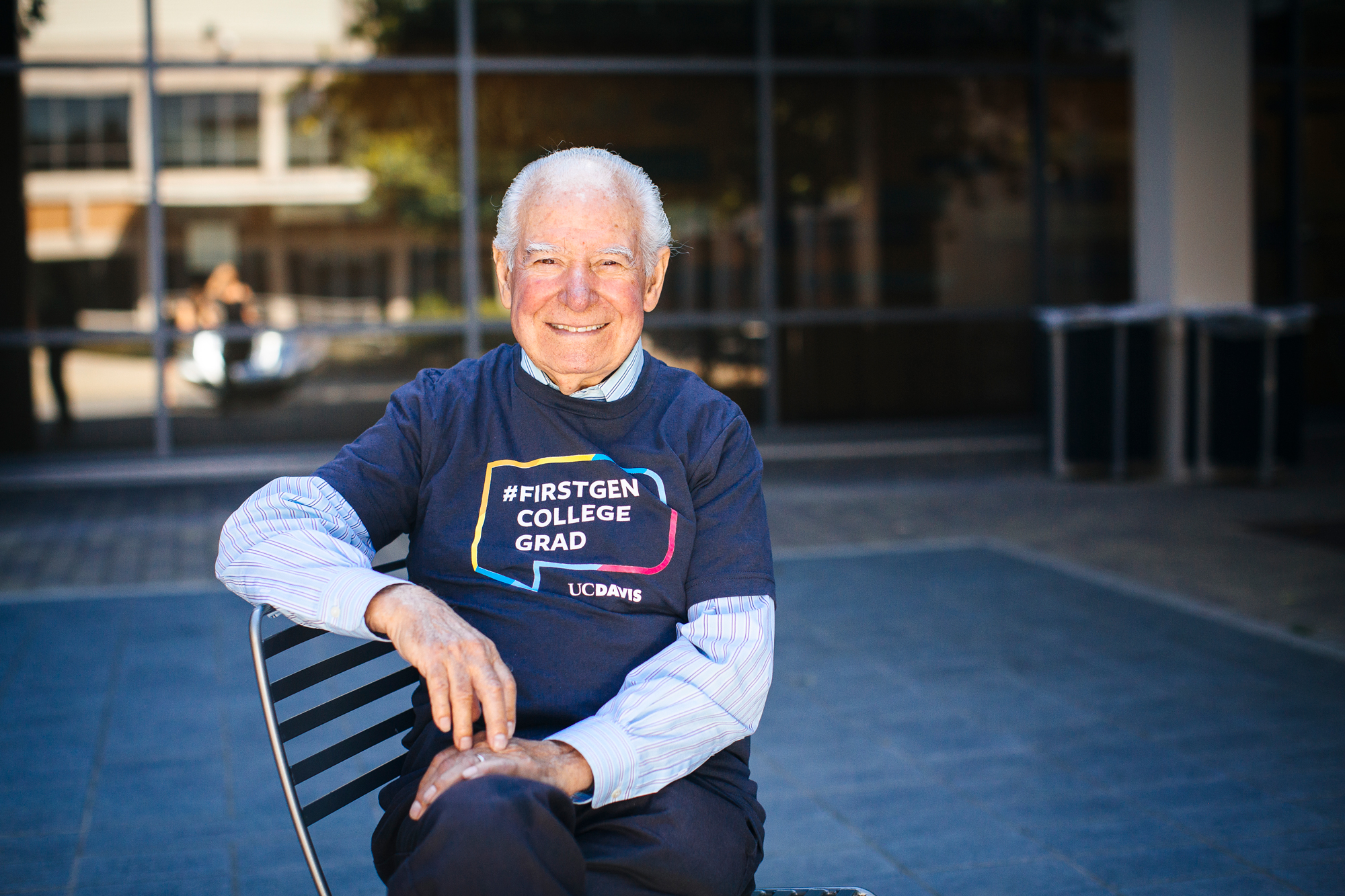 A man with gray hair in a first-generation college grad T-shirt sitting in front of a university building