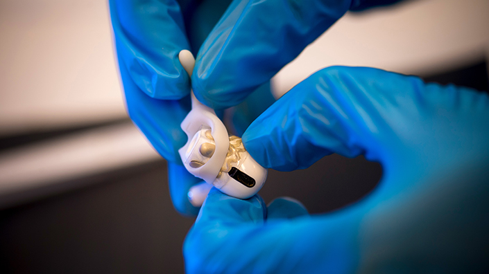 Blue gloved hands hold a white earbud with a silicon hook attached