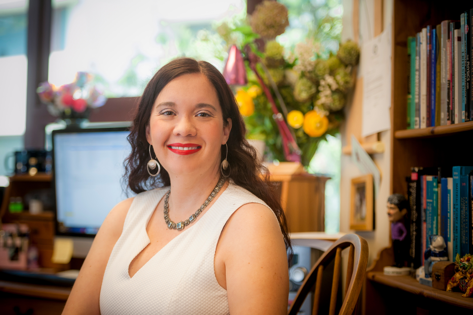 A smiling person sitting at a desk in an office with flowers and books