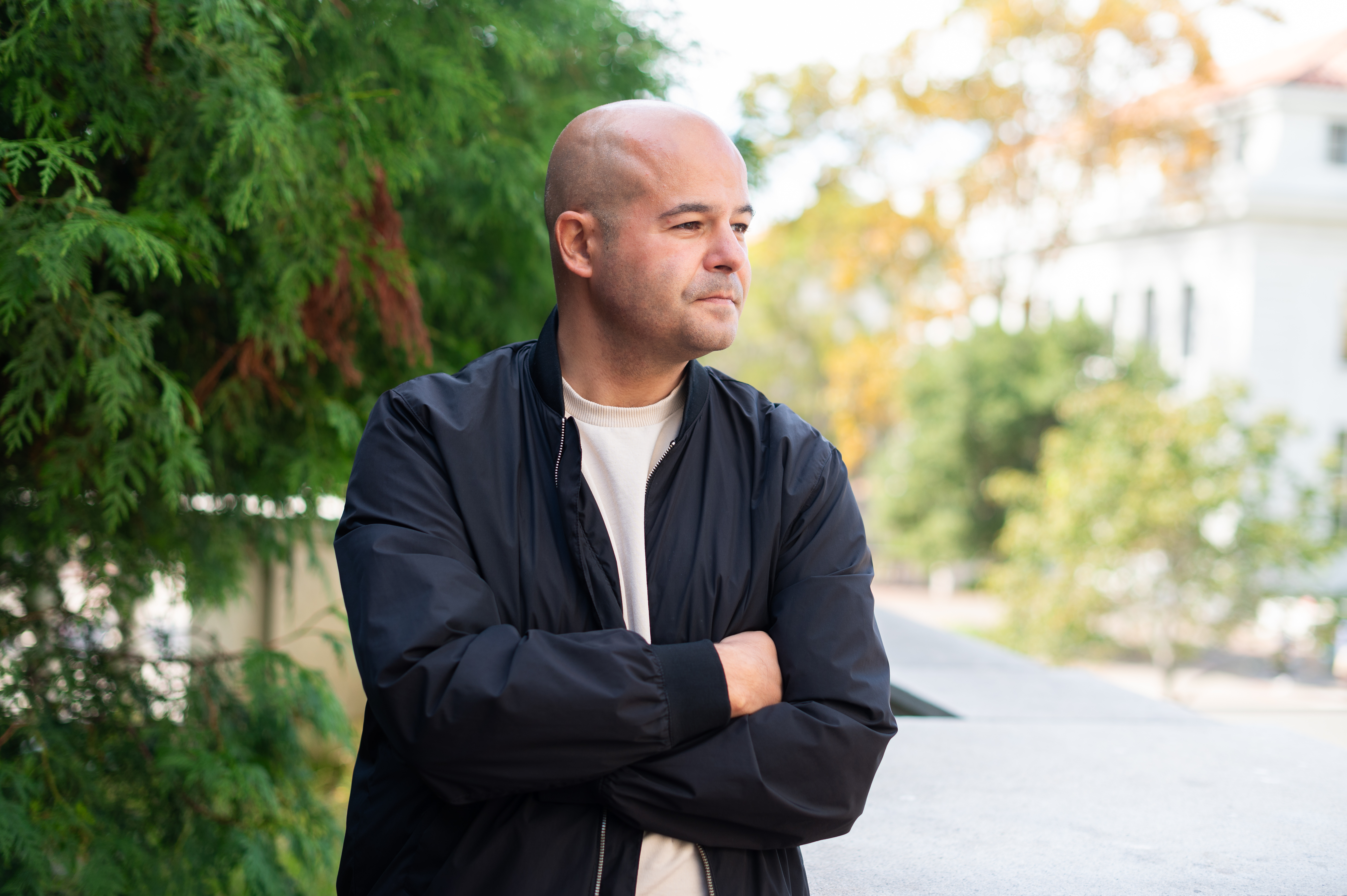 Ilya Budraitskis poses for a portrait looking away from the camera. Trees and a white building are visible in the background.