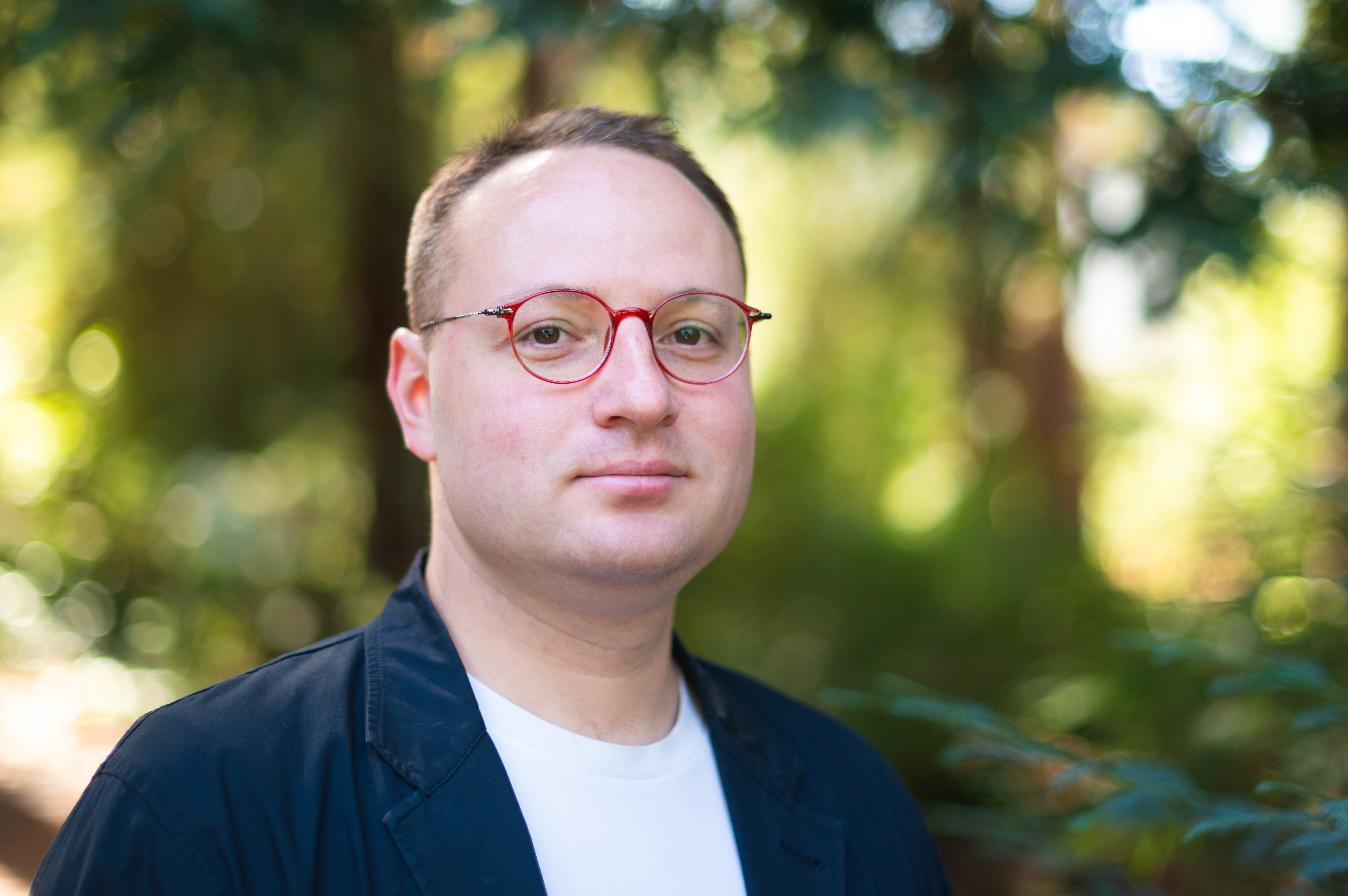 Ilya Matveev, wearing red-rimmed eyeglasses, faces the camera for a shoulders-up portrait against a background of outdoor greenery.