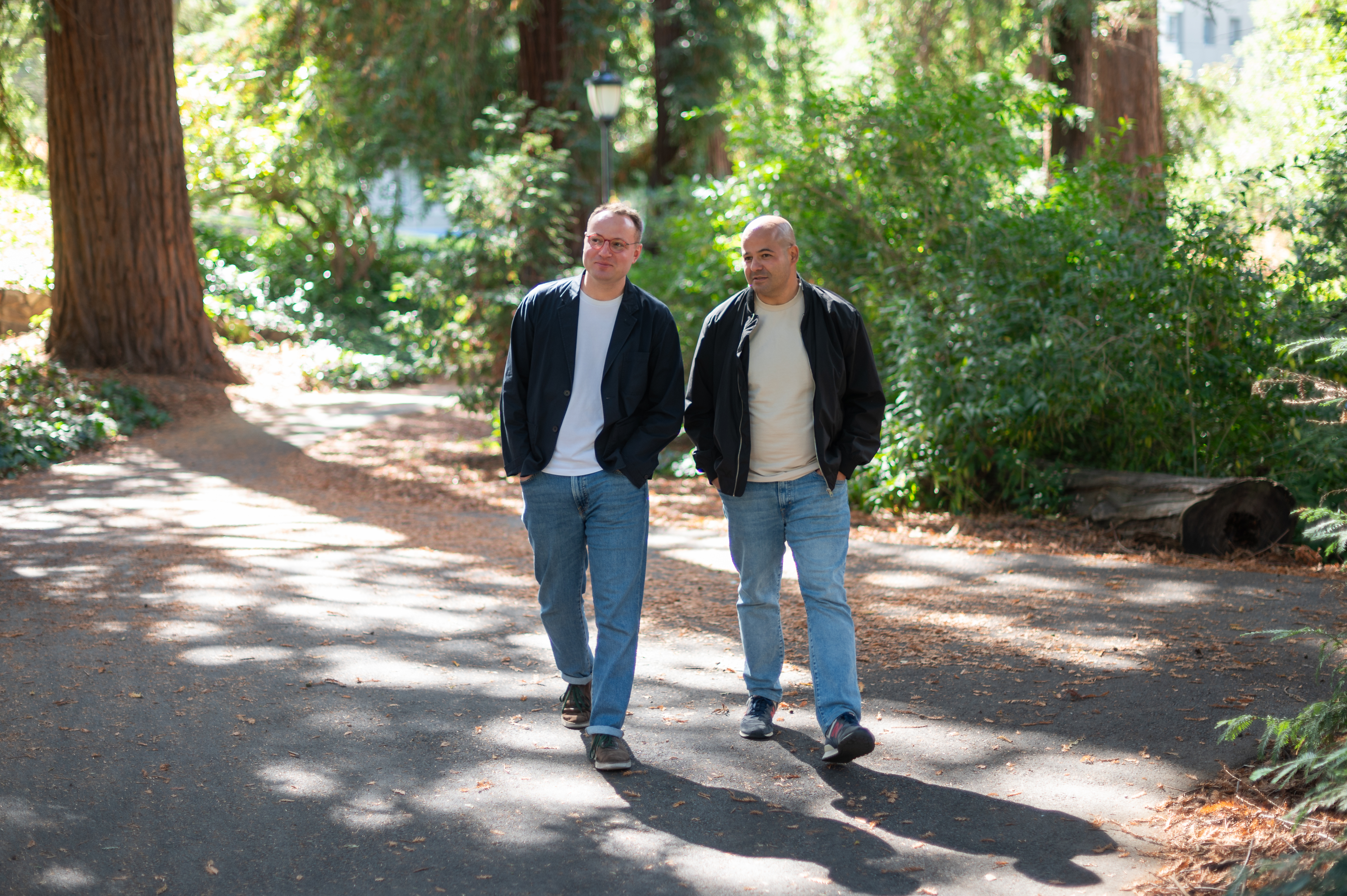 Two men walk toward the camera in a shady grove of redwoods on a paved pathway.
