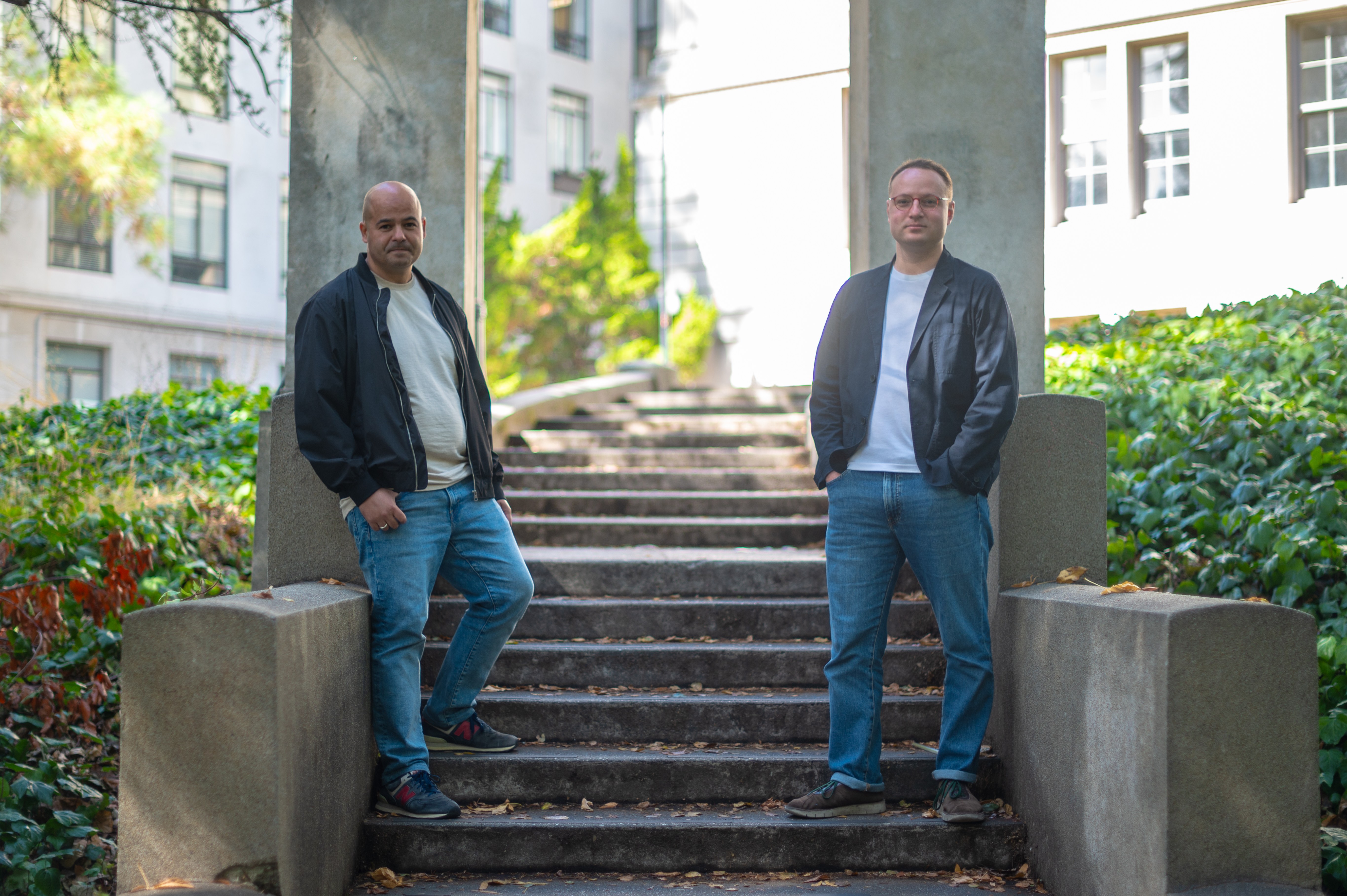 Two men in jeans and dark jackets pose for a portrait on an outdoor staircase