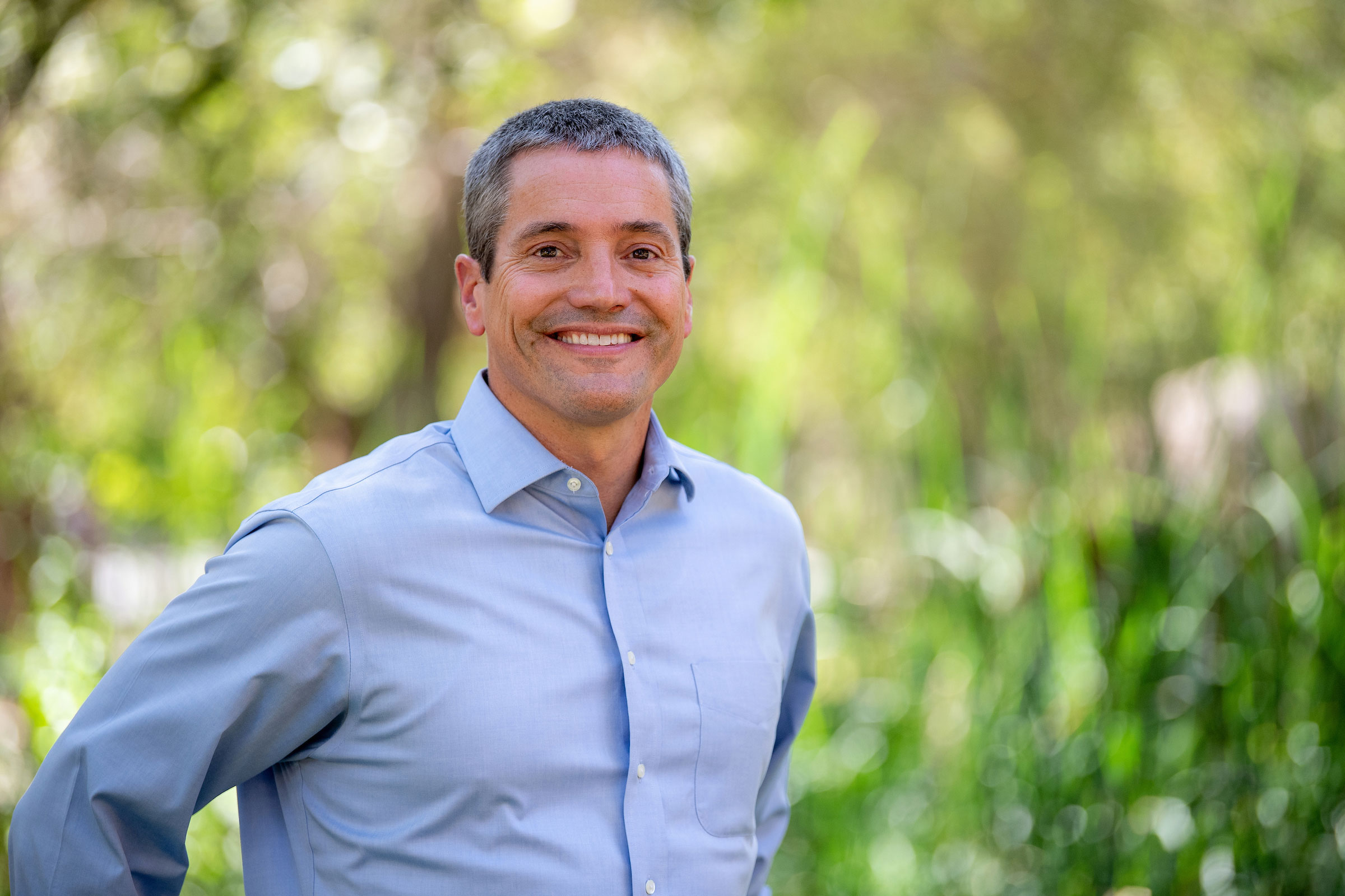 Wade Crowfoot, with close-cropped salt and pepper hair, wearing a blue button up shirt, smiles for a portrait with soft-focus greenery in the background.