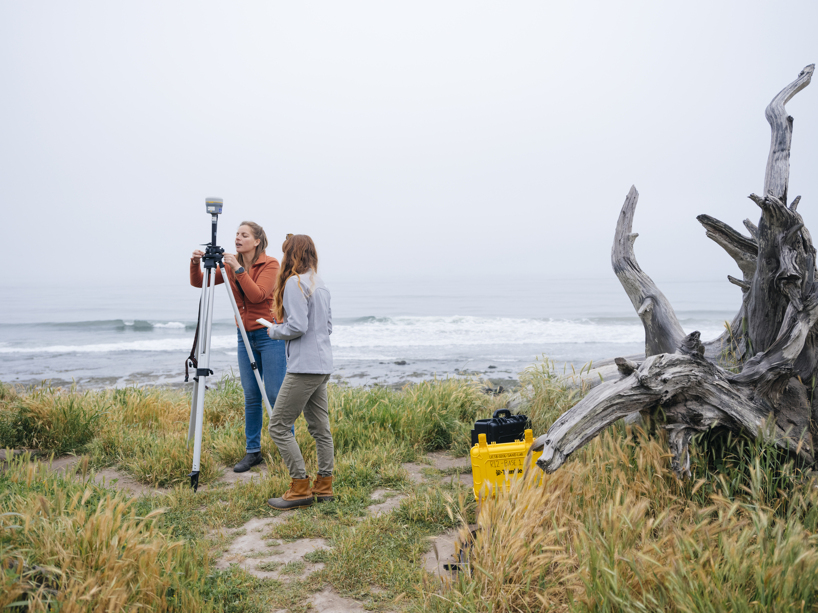 Two young women look into a small device on a tripod, which is set up on a grassy patch of land with breaking waves in the background.