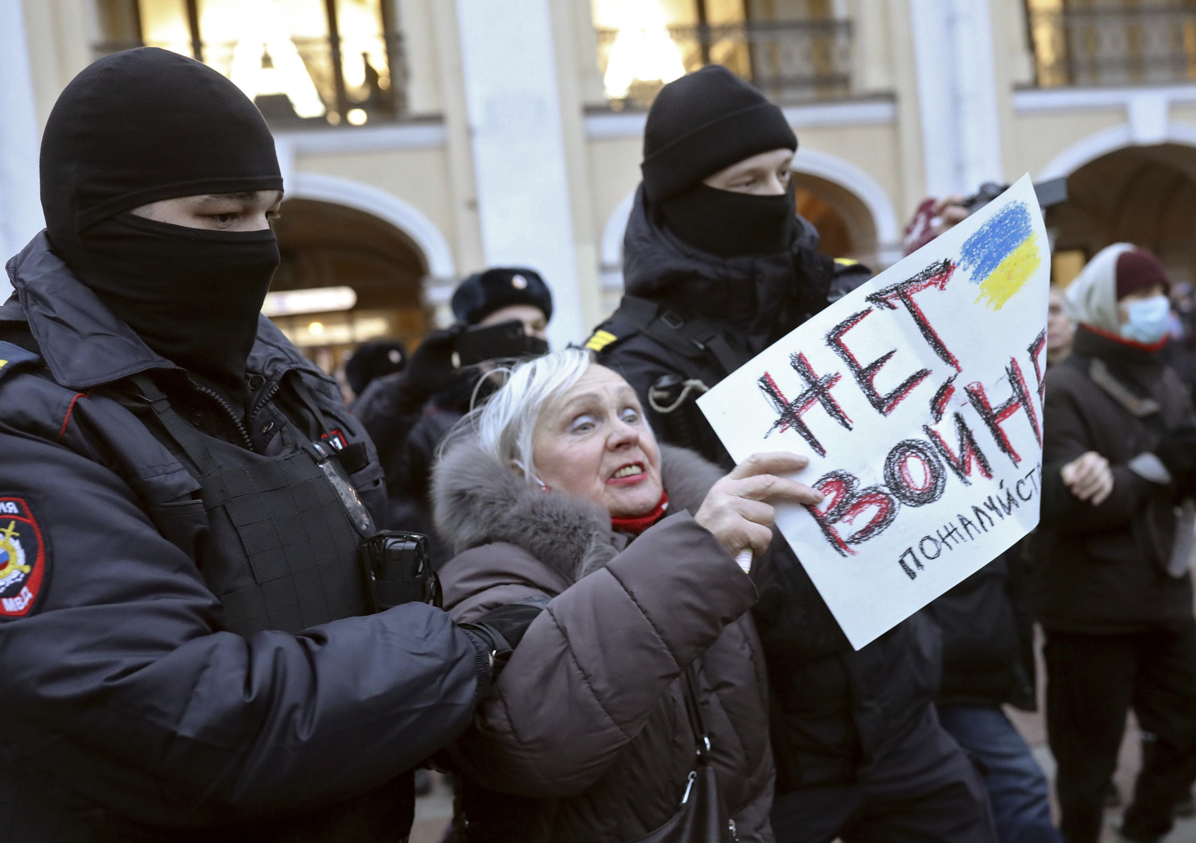 An older woman holding a sign in Russian is handled by police wearing balaclavas.
