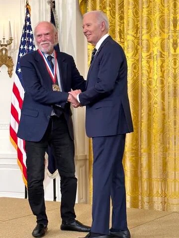 Barry Barish, a white man with white hair and beard, wearing a suit and a medal around his neck, shakes hands with President Biden.