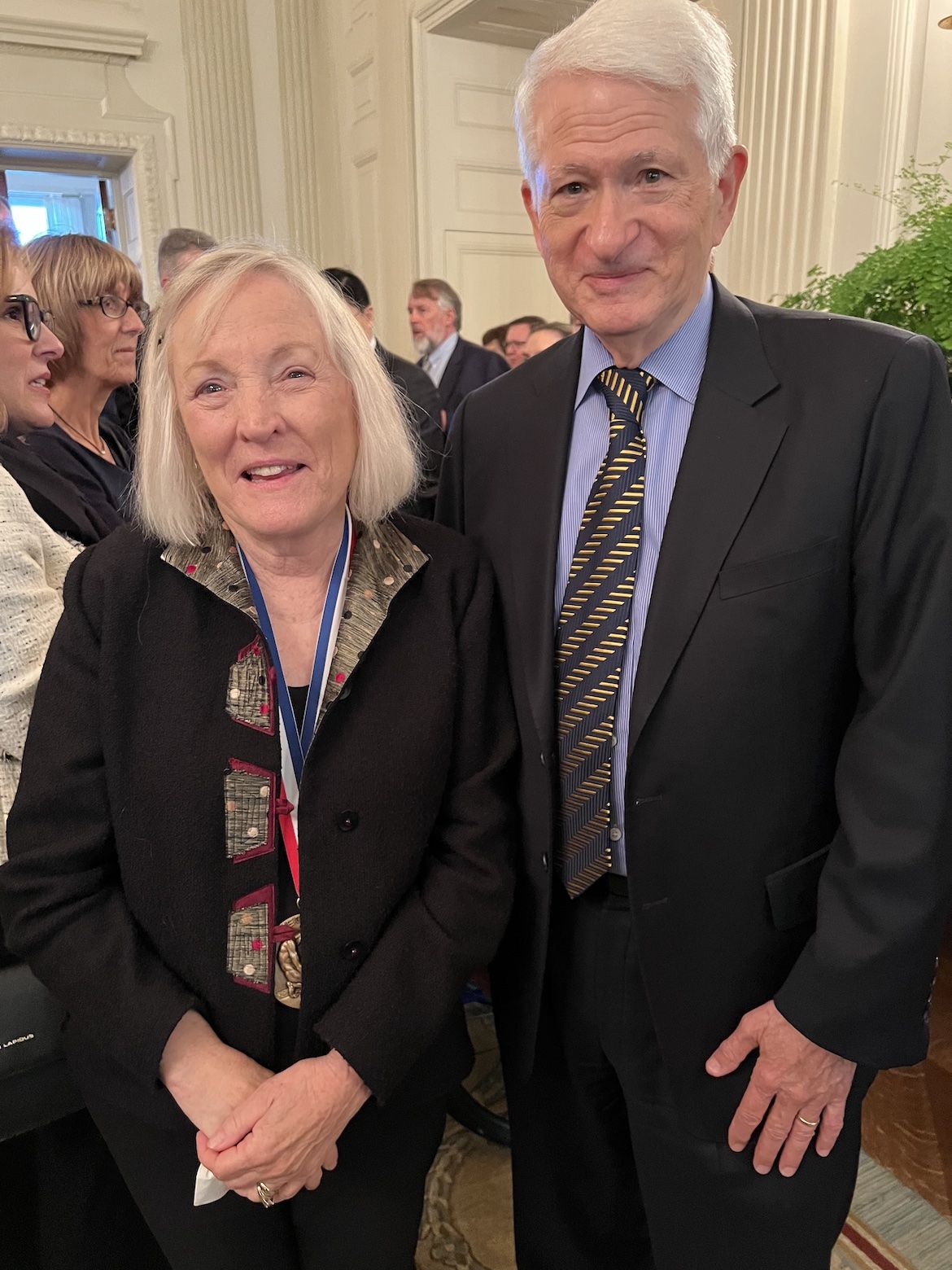 Shelley Taylor, white with a white/blonde bob haircut, wears a medal and smiles for the camera with UCLA Chancellor Gene Block, white with white hair, wearing a suit.