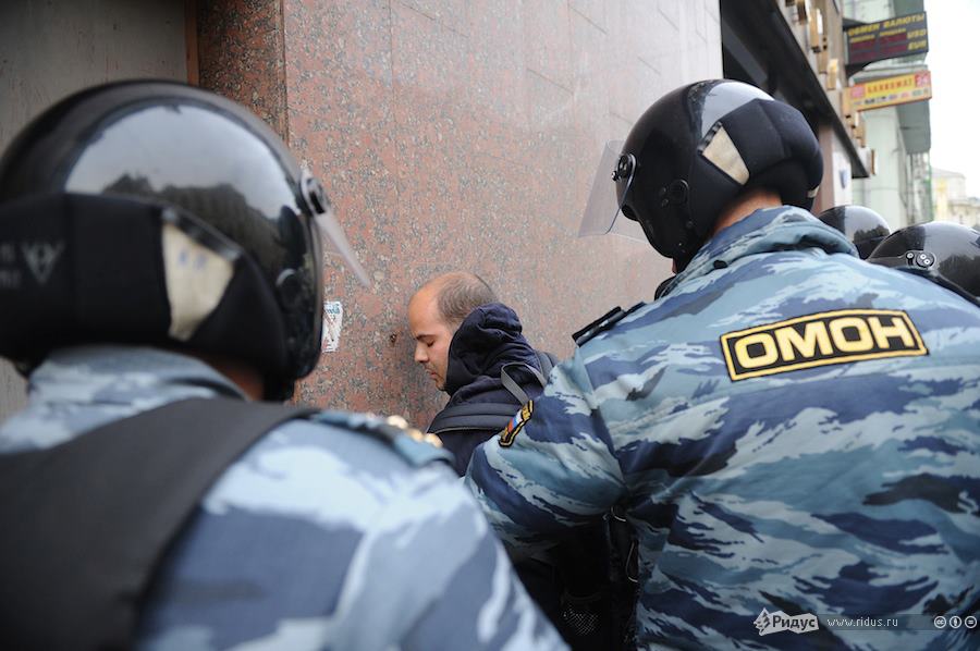 A man in a dark sweatshirt is held against a marble wall by two agents in camouflage and helmets. 