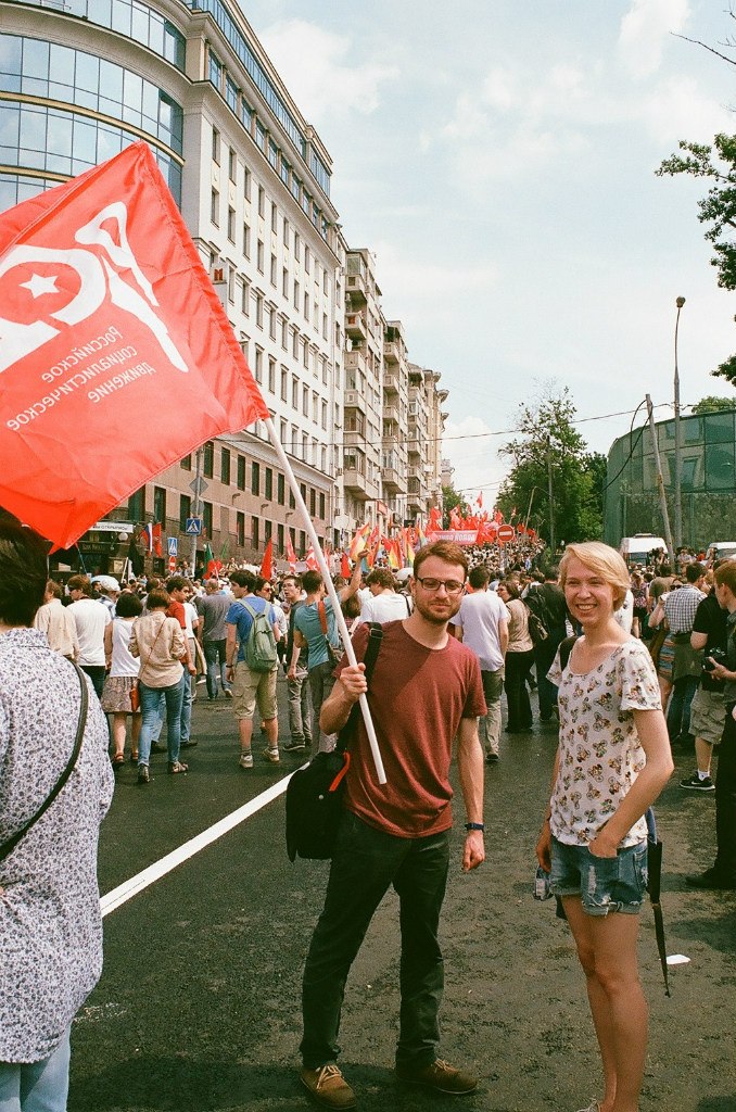 Ilya Matveev, wearing dark pants and a red shirt and waving a red flag, and a blonde woman wearing shorts and a t-shirt, at a rally