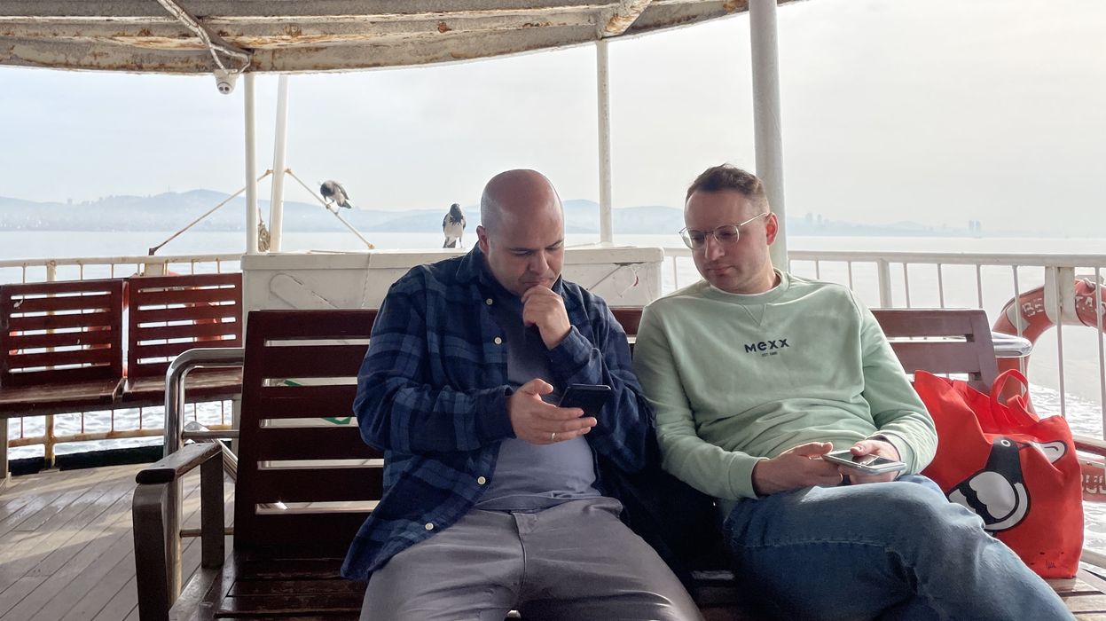 Two men sit on a bench, with the ocean visible in the background, looking at a phone, with focused expressions on their faces