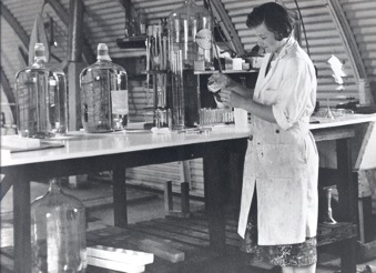 Black and white image of a brunette woman wearing a lab coat at a lab bench with glass instruments. 