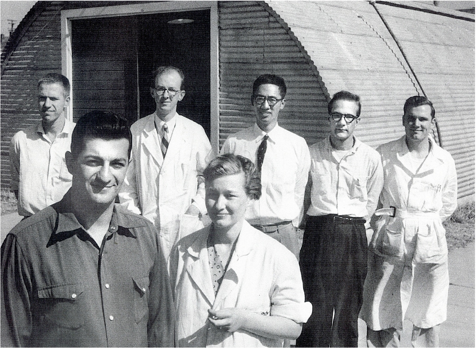 Black and white photograph of a young couple in the foreground, five people wearing lab coats in the middle ground, and a quonset hut in the background