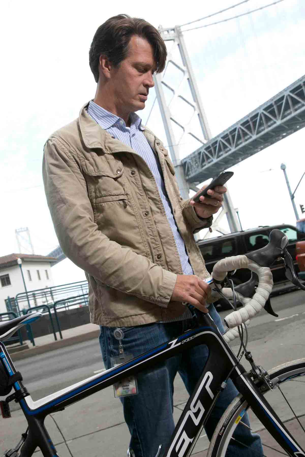 A brown-haired man looks at his phone while holding a road bike with the Bay Bridge in the background