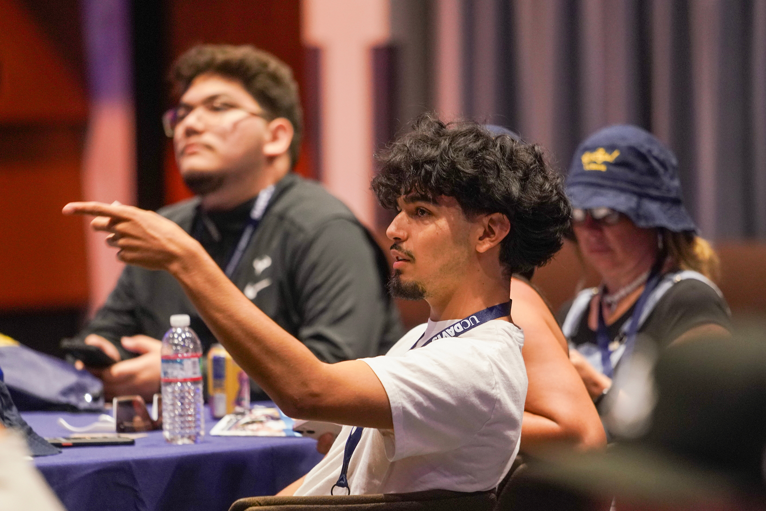 A student sitting at a table lifts his arm to point while asking a question